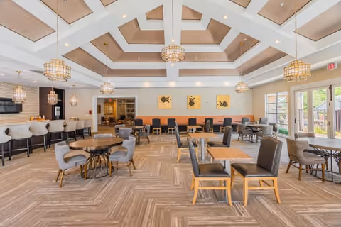 Spacious and well-lit dining area with multiple tables and chairs arranged neatly. The room features a high coffered ceiling with modern chandeliers, a bar area with stools on the left, and large windows and glass doors on the right allowing natural light to enter. The walls are decorated with three framed artworks above a long bench with cushions.