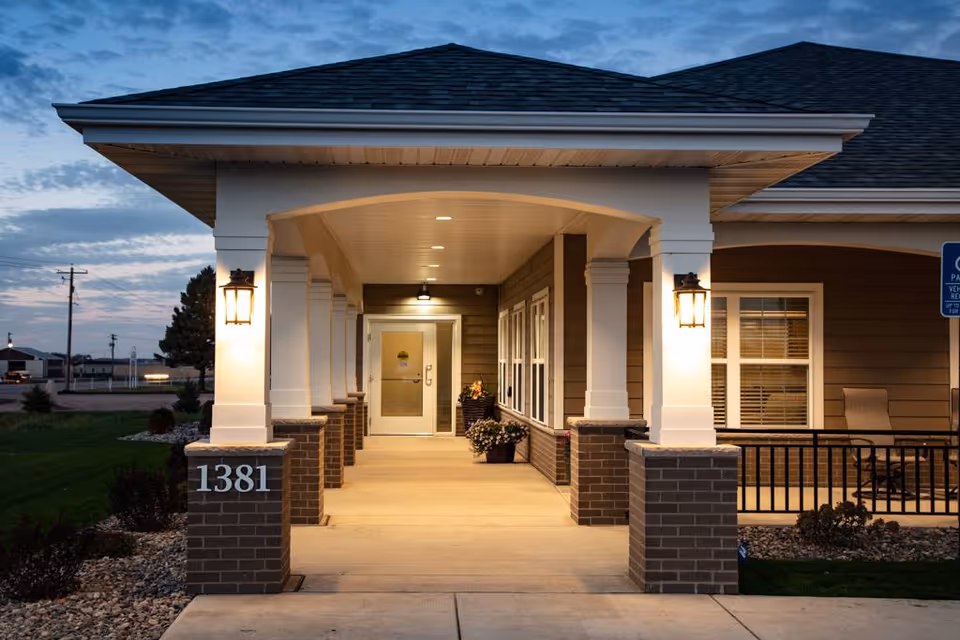 Entrance of a senior living facility at dusk with a covered walkway supported by white columns on brick bases, illuminated wall lanterns, potted plants near the door, and the building number 1381 displayed on one of the brick bases.