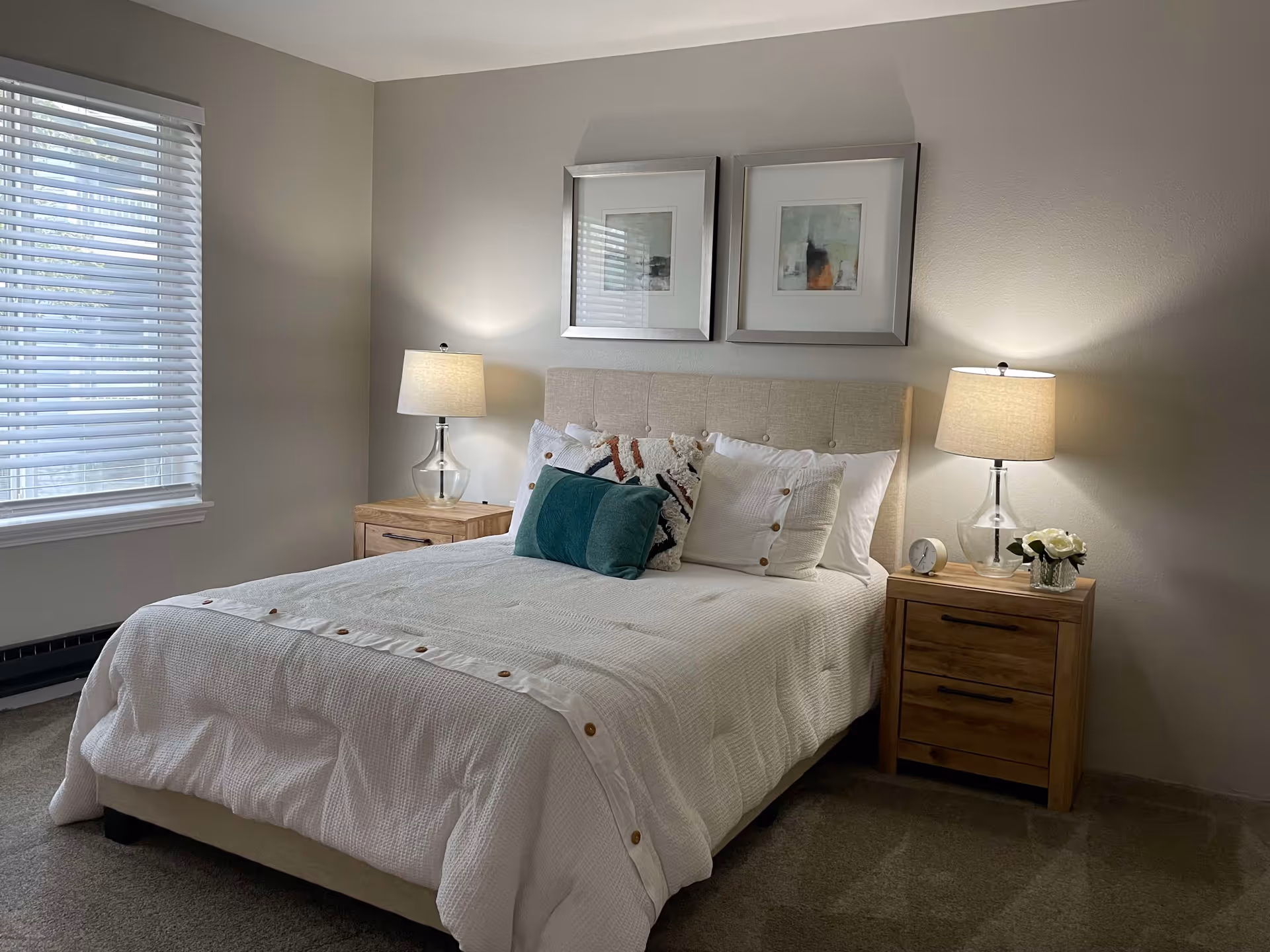 A neatly made bed with a beige upholstered headboard, white bedding with button details, and several decorative pillows including a teal one. On each side of the bed are wooden nightstands with glass-based lamps, a small clock, and a vase with white flowers. Two framed abstract artworks hang above the bed. A window with white blinds is on the left wall.