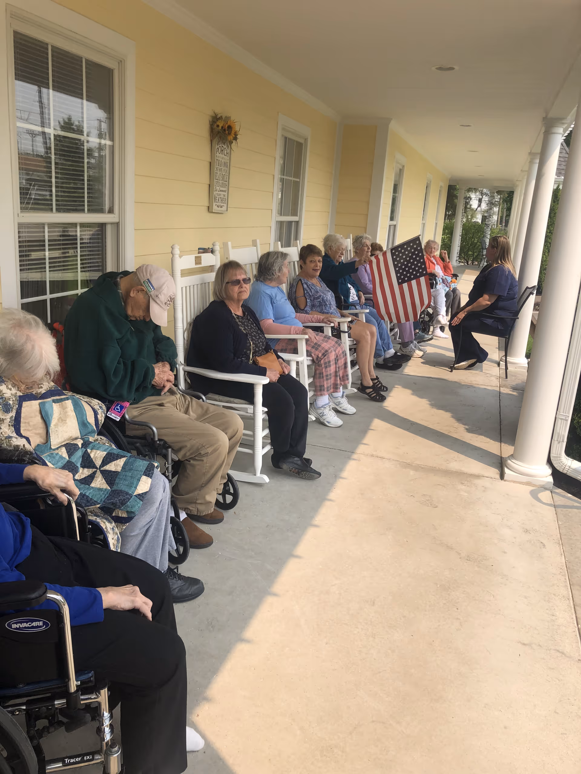 A group of elderly people sitting on rocking chairs and wheelchairs on a covered porch. One person is holding an American flag. A caregiver or staff member is seated at the far end of the porch. The porch has yellow siding, white columns, and windows along the wall.