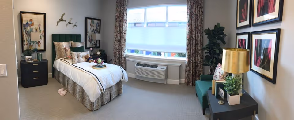 Sunlit furnished bedroom with a single bed, nightstands, framed artwork, an armchair, and a window air-conditioning unit.