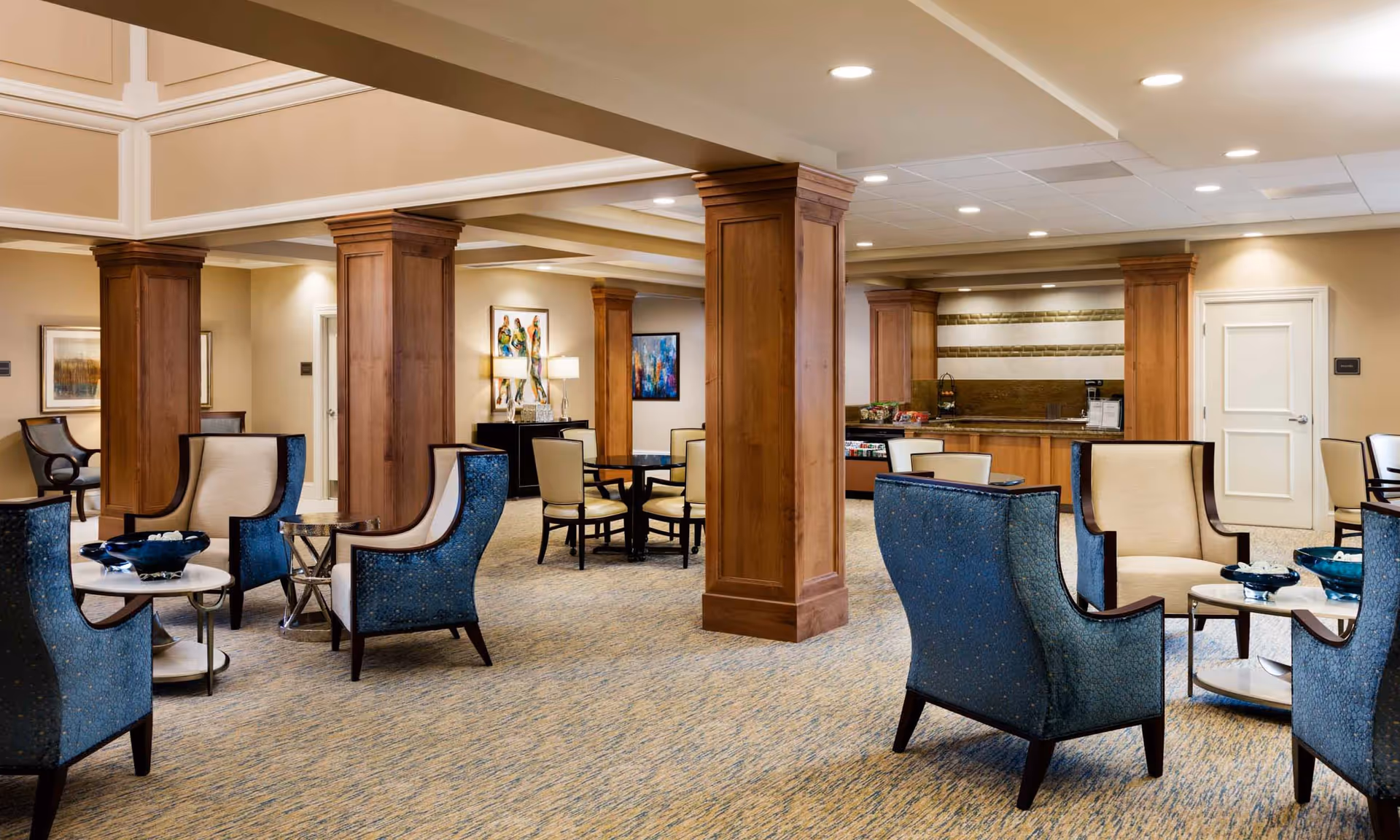 A spacious and well-lit senior living facility common area with beige walls and carpeted floors. The room features several wooden pillars, blue and beige upholstered armchairs arranged around small round tables, and a dining area with tables and chairs in the background. There is a counter area with snacks and a coffee machine, as well as framed artwork on the walls.