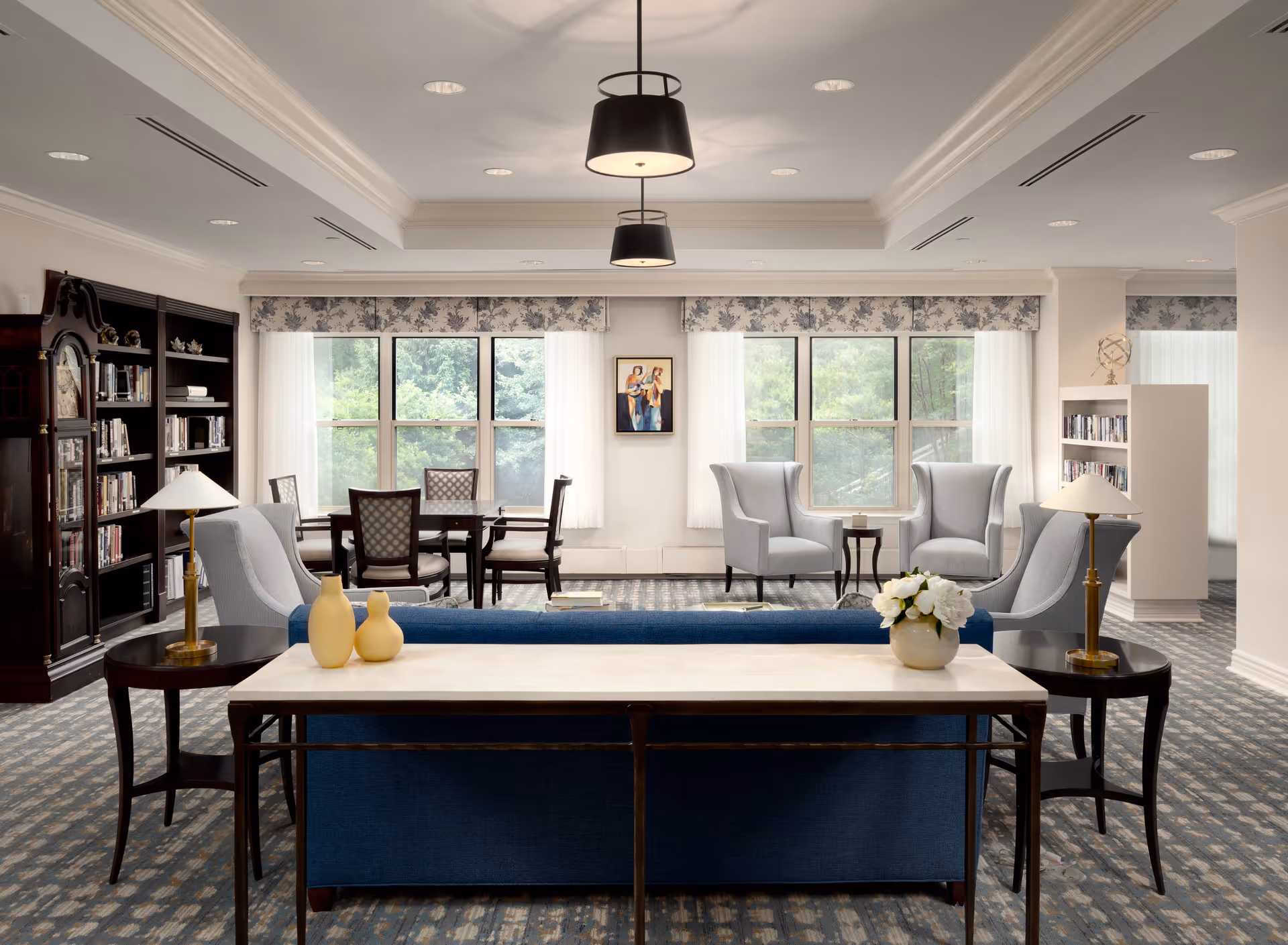 A bright and spacious living room area in a senior living facility with large windows letting in natural light. The room features a blue sofa with a marble-topped console table behind it, two light gray armchairs with a small round table between them, a wooden dining table with four chairs, bookshelves filled with books, and decorative lamps and vases. The walls are light-colored, and the carpet has a patterned design.