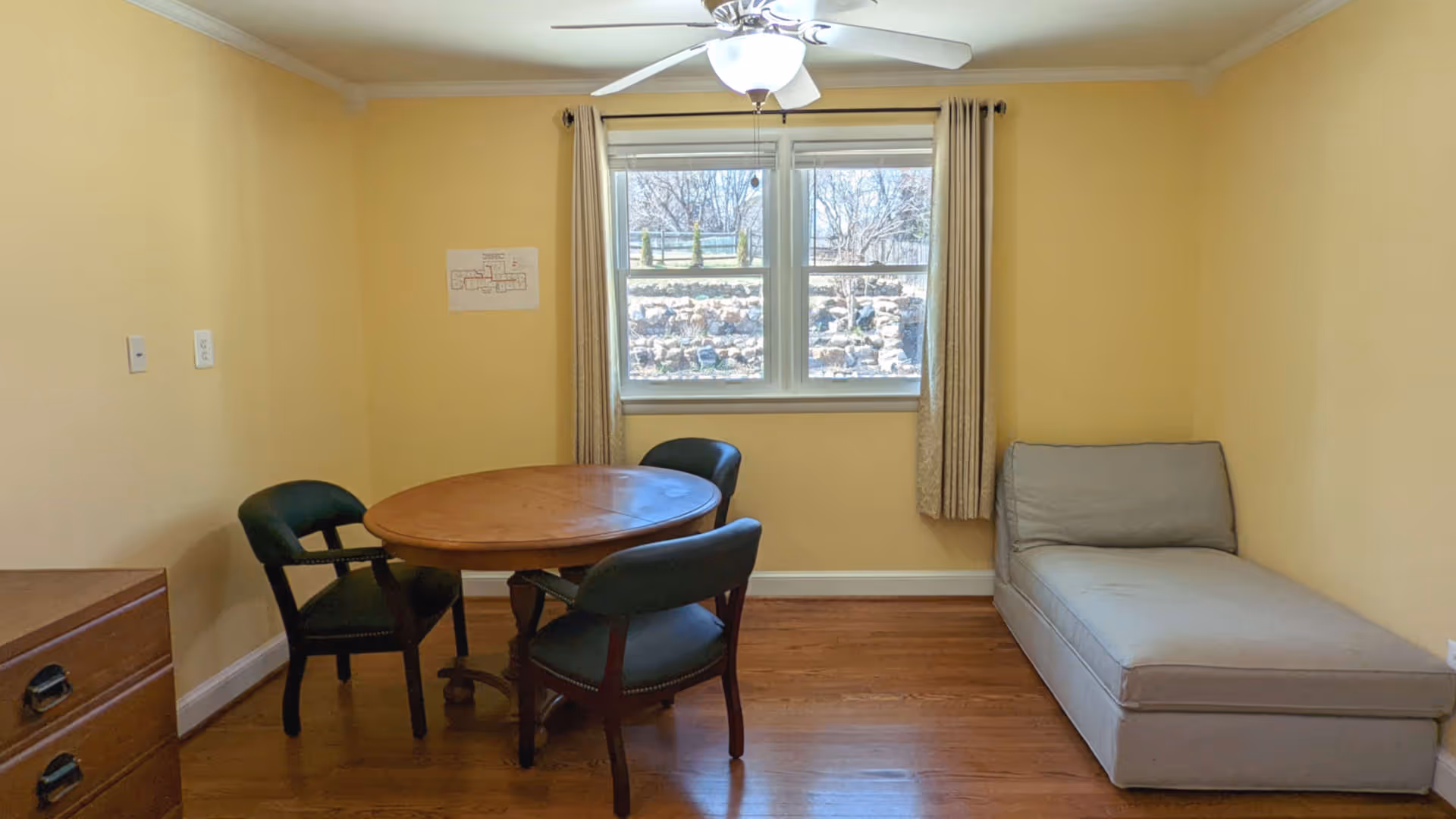 A small room with yellow walls and wooden flooring featuring a round wooden table surrounded by three dark green upholstered chairs. A light gray chaise lounge is positioned against the right wall. A window with beige curtains lets in natural light, and a ceiling fan with a light fixture is mounted above.