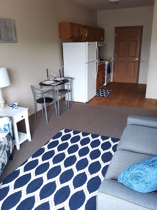 View of a senior living apartment interior showing a small kitchen area with wooden cabinets, a white refrigerator, and a stove. In front of the kitchen is a small round dining table set for two with blue candles and wine glasses. The living area has a gray couch with a blue pillow and a blue and white patterned rug. A white side table with a lamp and decorative bird is next to a floral-patterned chair.