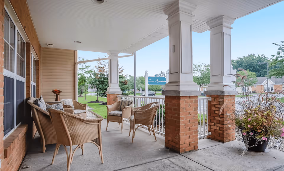 Covered outdoor patio area with wicker chairs and a small table, surrounded by brick pillars and white railings. There is a large planter with flowers on the right side and a green lawn with trees in the background under a clear blue sky.