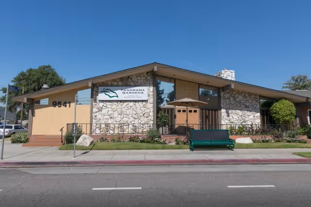 Exterior front view of Panorama Gardens Nursing & Rehabilitation Center, a single-story building with stone and wood paneling, large windows, a green bench, and a sign displaying the facility's name.