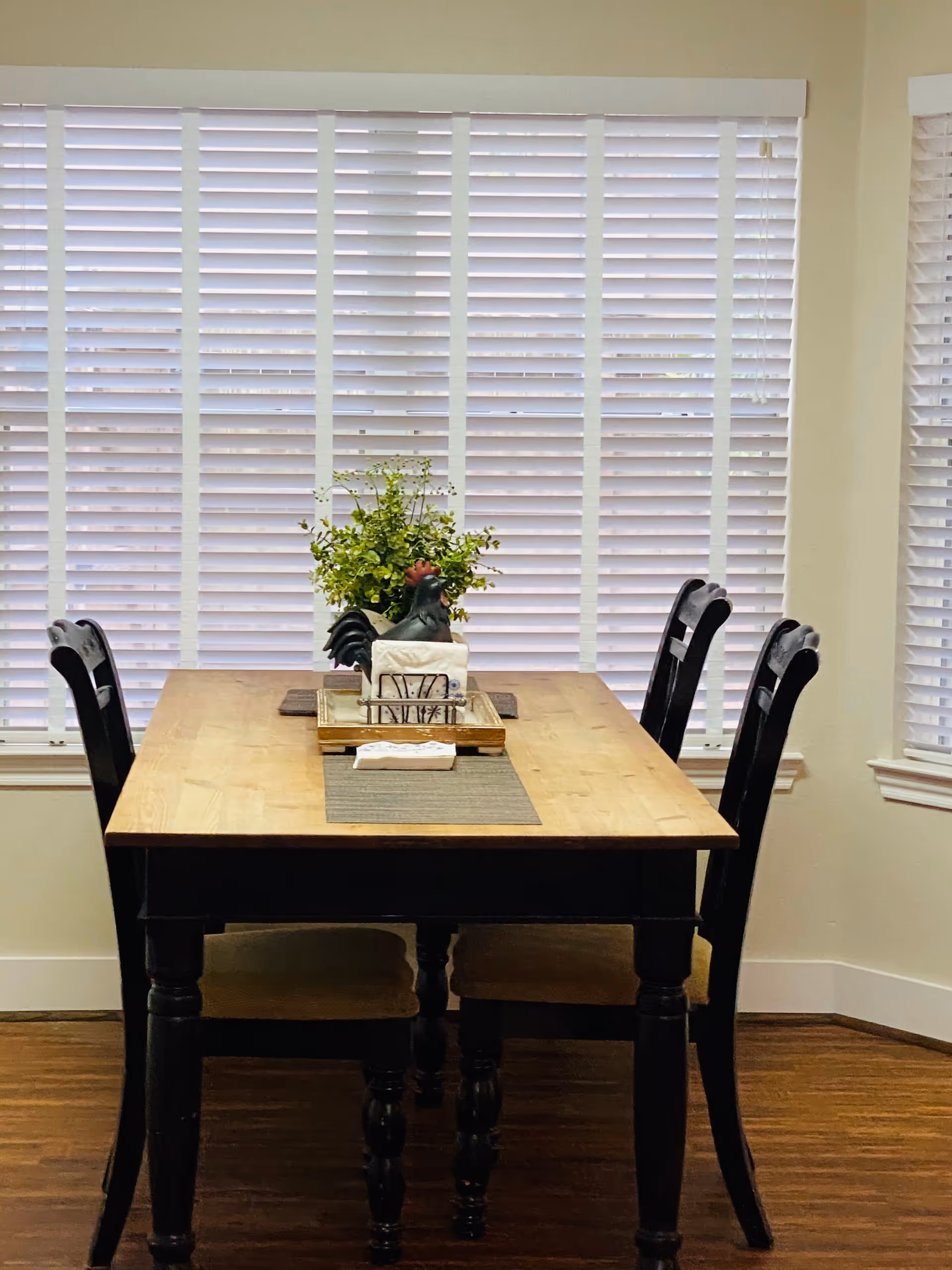 A wooden dining table with four black chairs around it, placed in front of large windows with white blinds. On the table is a centerpiece with a green plant and a decorative rooster figure, along with a napkin holder and placemats.