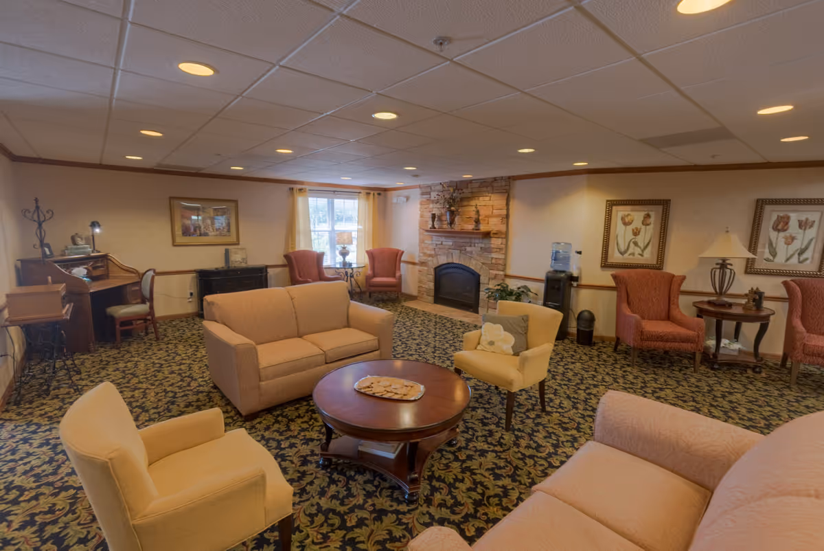 A cozy living room with patterned carpet, beige and red upholstered chairs and sofas arranged around a wooden coffee table with a tray of cookies. The room features a stone fireplace with decorative items on the mantel, framed floral artwork on the walls, a water cooler, and a small wooden desk with a chair near a window with yellow curtains.