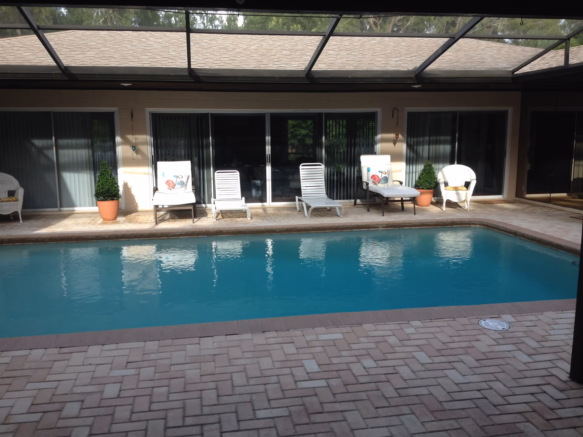 Outdoor swimming pool with clear blue water surrounded by a brick patio. Several lounge chairs and potted plants are arranged along the wall of a building with sliding glass doors. The area is covered by a screened enclosure.