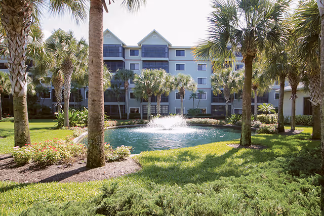 A landscaped outdoor area at Harbour Health Assisted Living featuring a small pond with a water fountain in the center, surrounded by palm trees, green grass, and flowering plants. In the background, there is a multi-story residential building with balconies and screened-in porches.