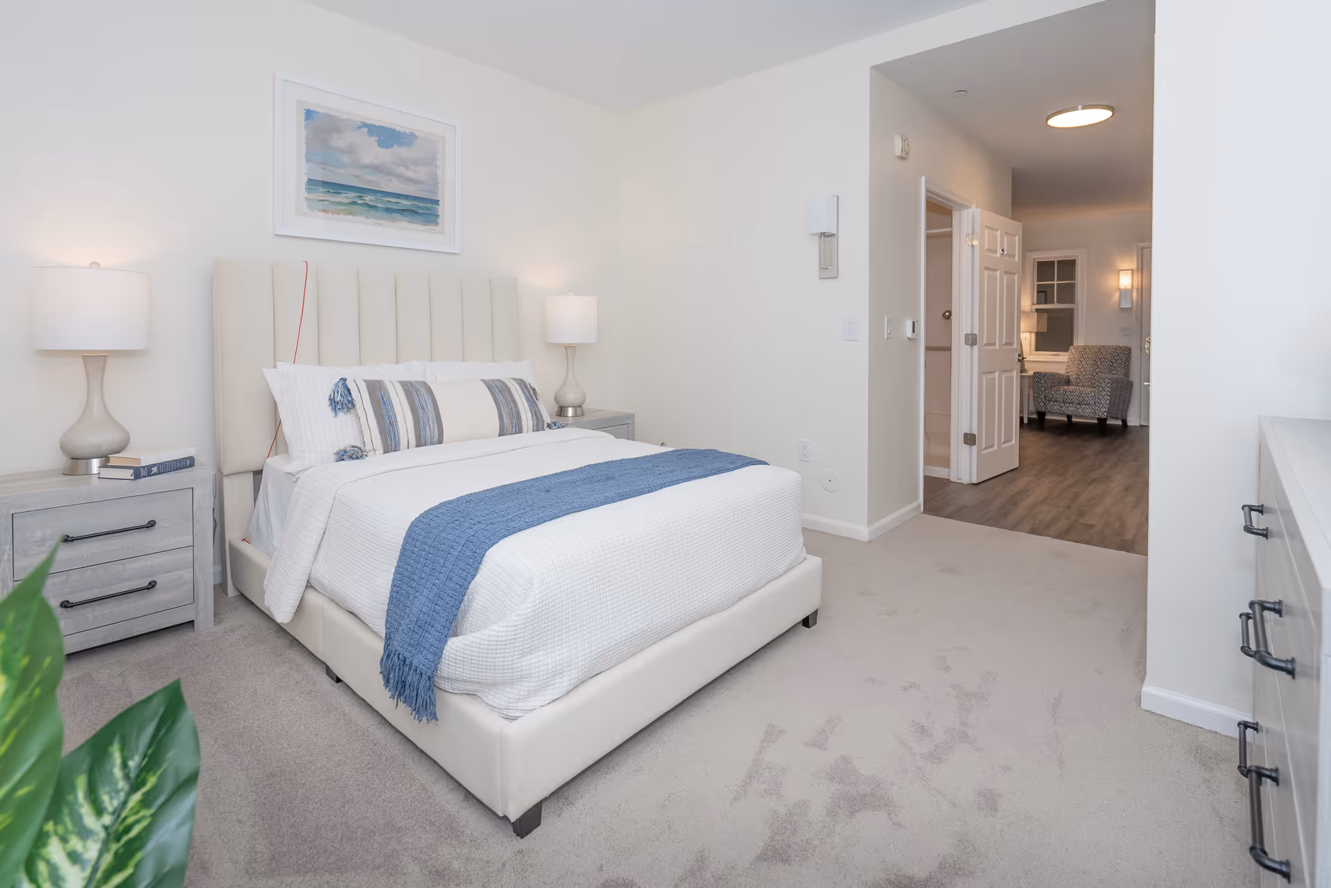 A bright and clean bedroom with a white upholstered bed featuring white bedding and a blue throw blanket. There are two matching bedside tables with lamps on each side of the bed. A framed picture of a seascape hangs above the bed. The room has light-colored carpet flooring and white walls. An open doorway leads to another room with wood flooring and a patterned armchair.