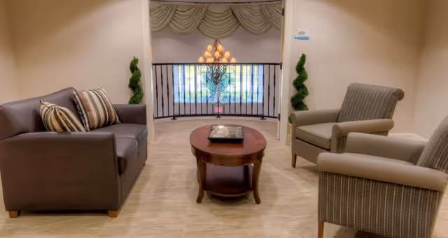 A cozy seating area with a brown sofa adorned with striped pillows on the left, two beige armchairs on the right, and a wooden coffee table in the center. In the background, there is a decorative chandelier hanging in front of a window with an ornate wrought iron railing and draped curtains.