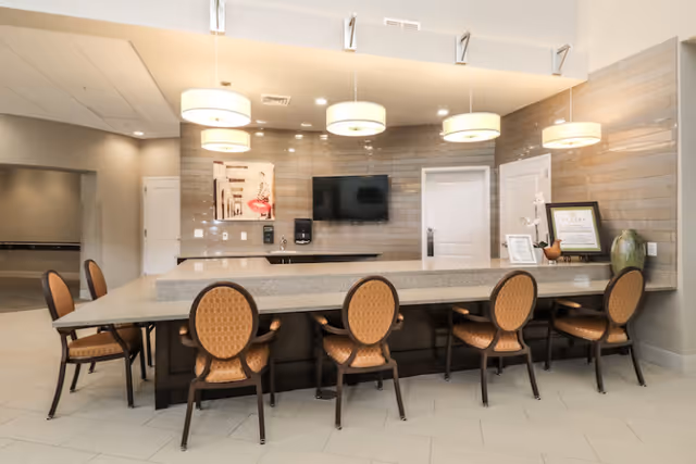 Bright communal interior with a long countertop and six upholstered chairs facing a wall-mounted TV and pendant lights.
