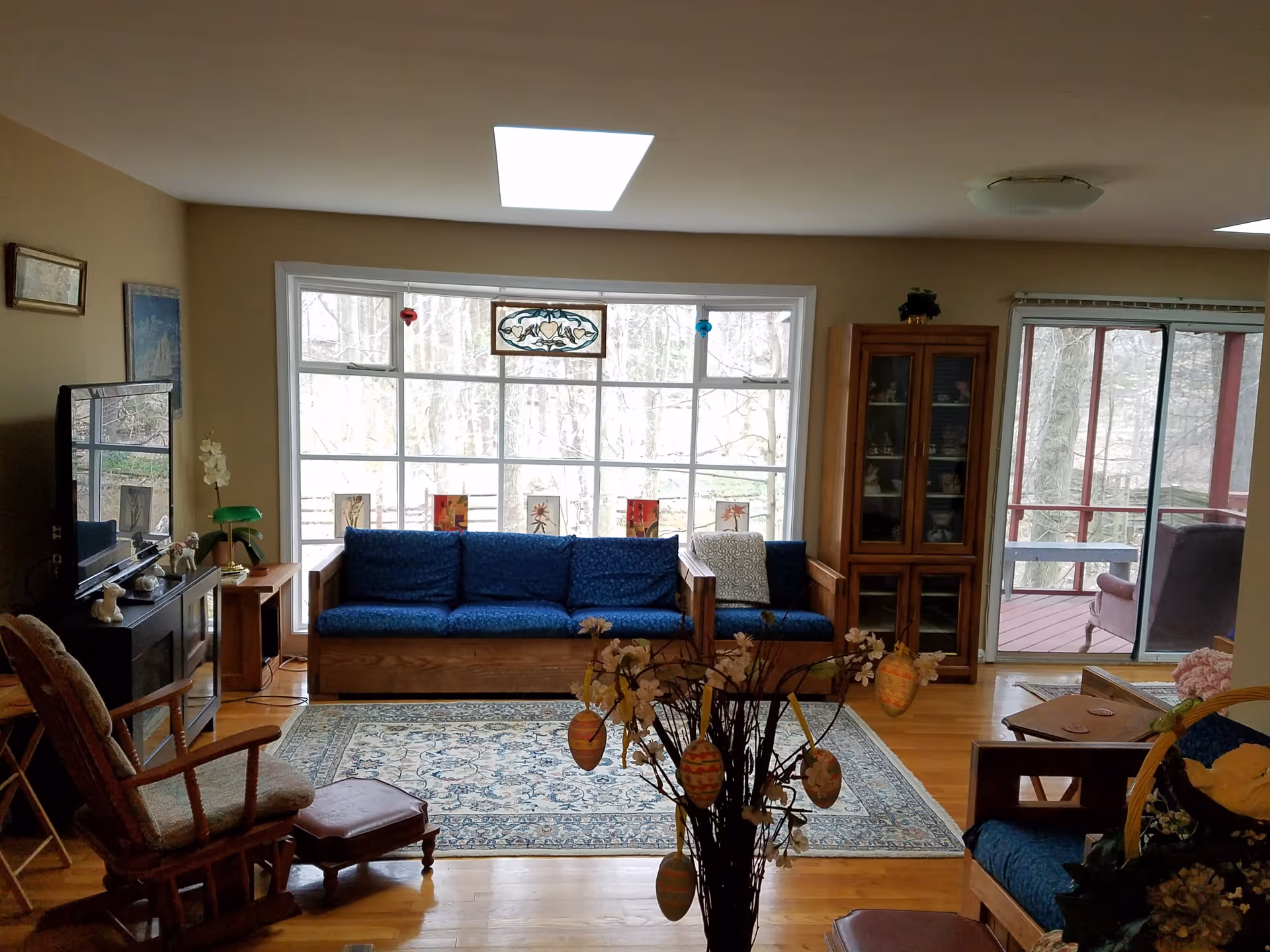 A cozy living room with wooden furniture including a blue cushioned sofa, a rocking chair with a footstool, and a glass-front cabinet. Large windows and a sliding glass door provide a view of trees outside. A patterned area rug covers part of the wooden floor, and decorative items hang from a branch centerpiece in the foreground.