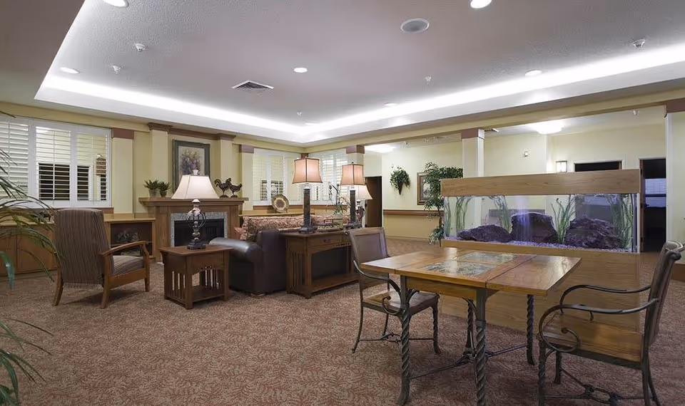 A cozy living room area in McLoughlin Place featuring a fireplace with decorative items on the mantel, a sofa, armchairs, side tables with lamps, a wooden table with chairs, and a large aquarium divider. The room has carpeted flooring, soft lighting, and plants adding to the warm ambiance.