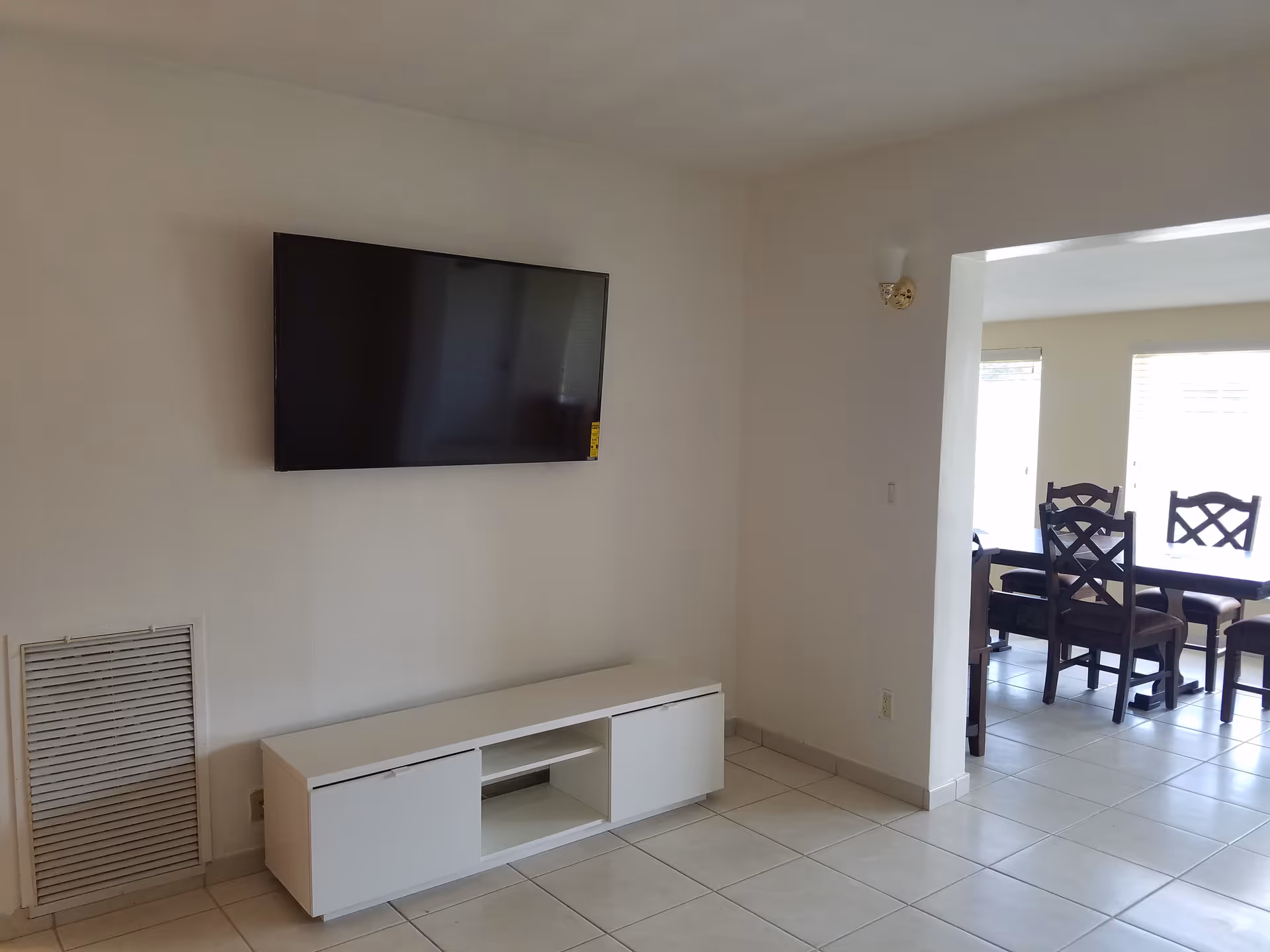 Interior view of a living area with a wall-mounted flat-screen TV above a white media console. Adjacent to this space is a dining area with a dark wooden table and chairs, illuminated by natural light from windows.