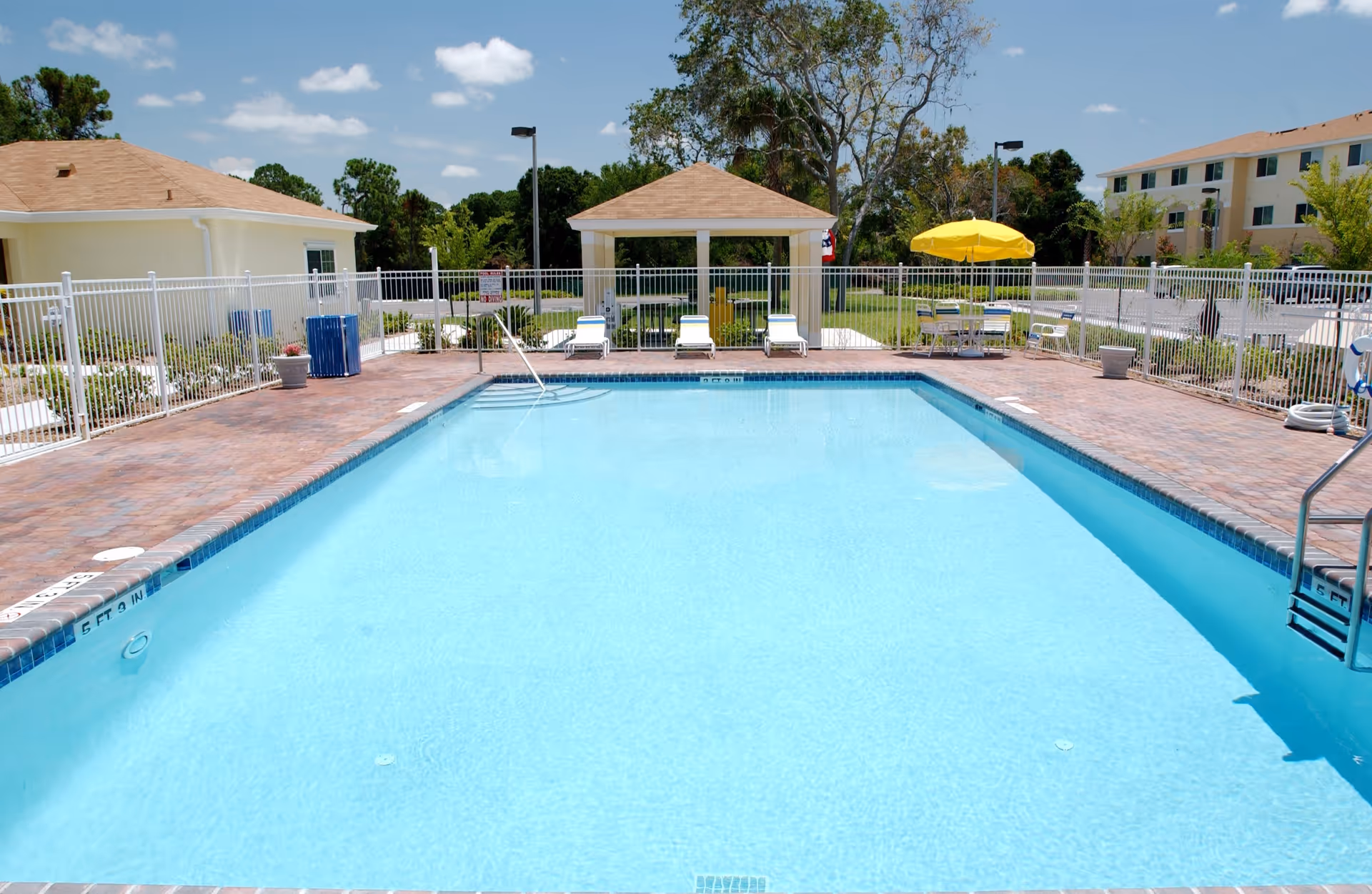 Outdoor swimming pool with clear blue water surrounded by a brick-paved deck. There are white lounge chairs and a small pavilion with a peaked roof at the far end of the pool. A yellow umbrella shades a table with chairs nearby. The area is enclosed by a white metal fence, and there are buildings and trees in the background under a partly cloudy sky.