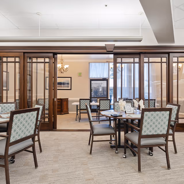 A bright dining room in a retirement community with multiple tables and chairs arranged neatly. The chairs have a patterned backrest and cushioned seats. The room features large wooden framed glass partitions, beige carpet, and soft lighting from chandeliers. A window with sheer curtains allows natural light to enter the space.