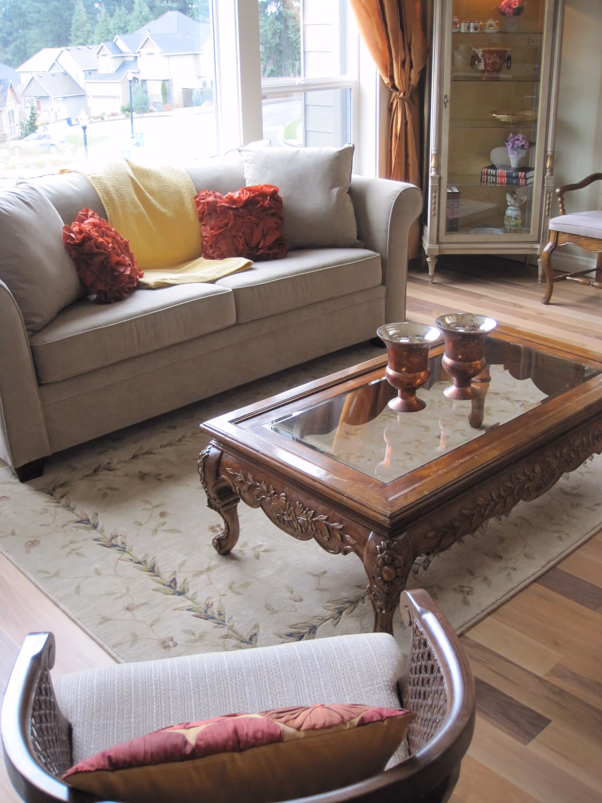 A cozy living room with a beige sofa adorned with red and beige pillows and a yellow throw blanket. In front of the sofa is an ornate wooden coffee table with a glass top, holding two decorative vases. A cushioned chair with a red and beige pillow is partially visible in the foreground. The room has wooden flooring, a patterned area rug, a glass display cabinet with decorative items, and large windows with brown curtains letting in natural light.