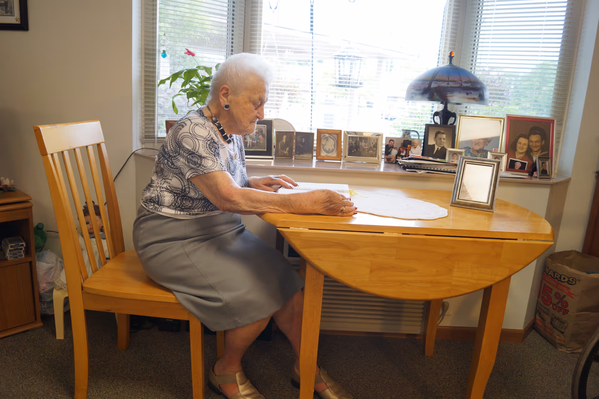 An elderly woman with white hair sits at a wooden table in a room with a window behind her. The table has a white doily and a picture frame on it. Behind the table on the windowsill are multiple framed photographs and a decorative lamp. The woman appears to be engaged in an activity at the table. The room has a cozy, home-like atmosphere.