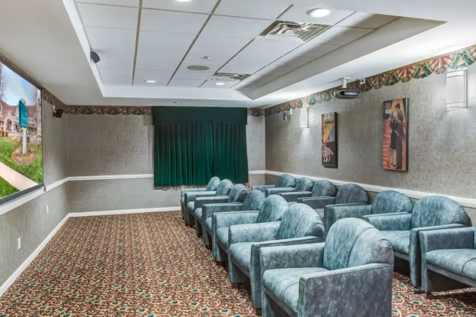 A small theater room with two rows of green upholstered armchairs facing a large screen on the left wall. The room has patterned carpet, gray walls with decorative border near the ceiling, two framed posters on the right wall, and green curtains covering a window at the back.