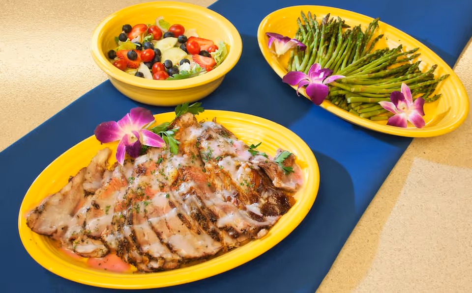 Three yellow plates on a blue table runner with food: a plate of sliced roasted meat with sauce and a purple flower garnish, a plate of steamed asparagus with purple flower garnishes, and a bowl of mixed fruit salad with blueberries, strawberries, grapes, and melon pieces.