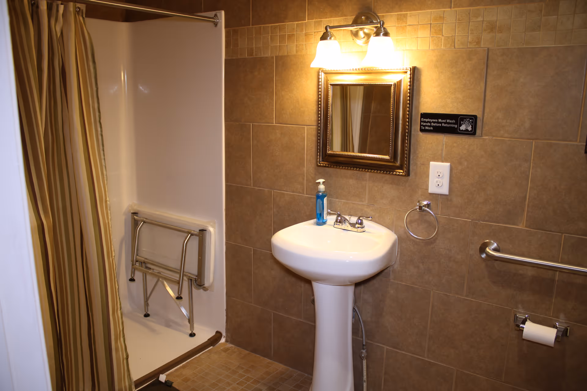 A bathroom with beige tiled walls and floor, featuring a pedestal sink with a soap dispenser, a mirror with a decorative frame above the sink, a light fixture with two bulbs, a shower area with a striped curtain and a foldable shower seat, and safety grab bars near the toilet paper holder.