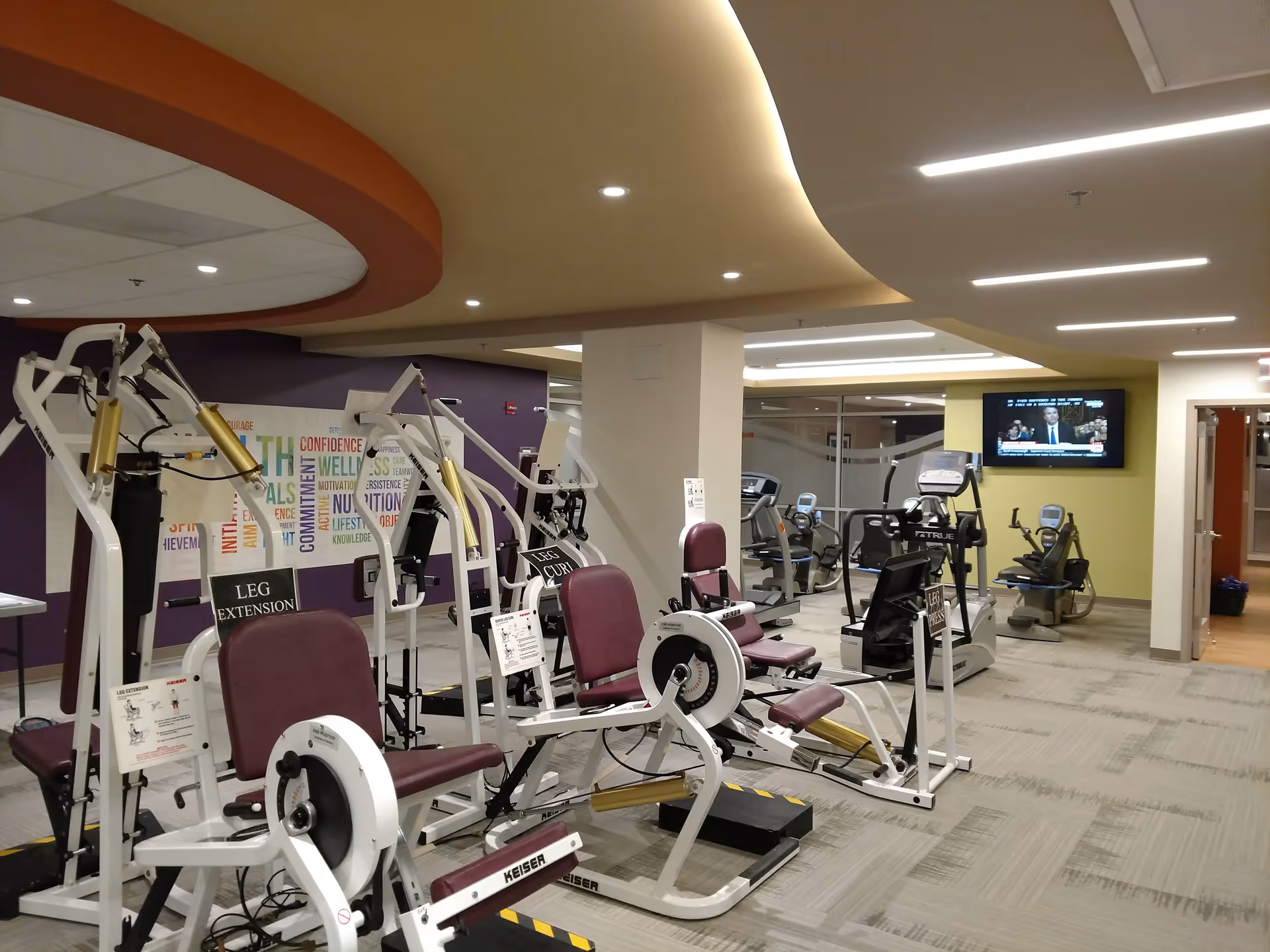 Interior view of a fitness center with various exercise machines including leg extension and leg curl equipment. The room has a modern design with a curved ceiling, a wall-mounted TV, and motivational word art on the wall.