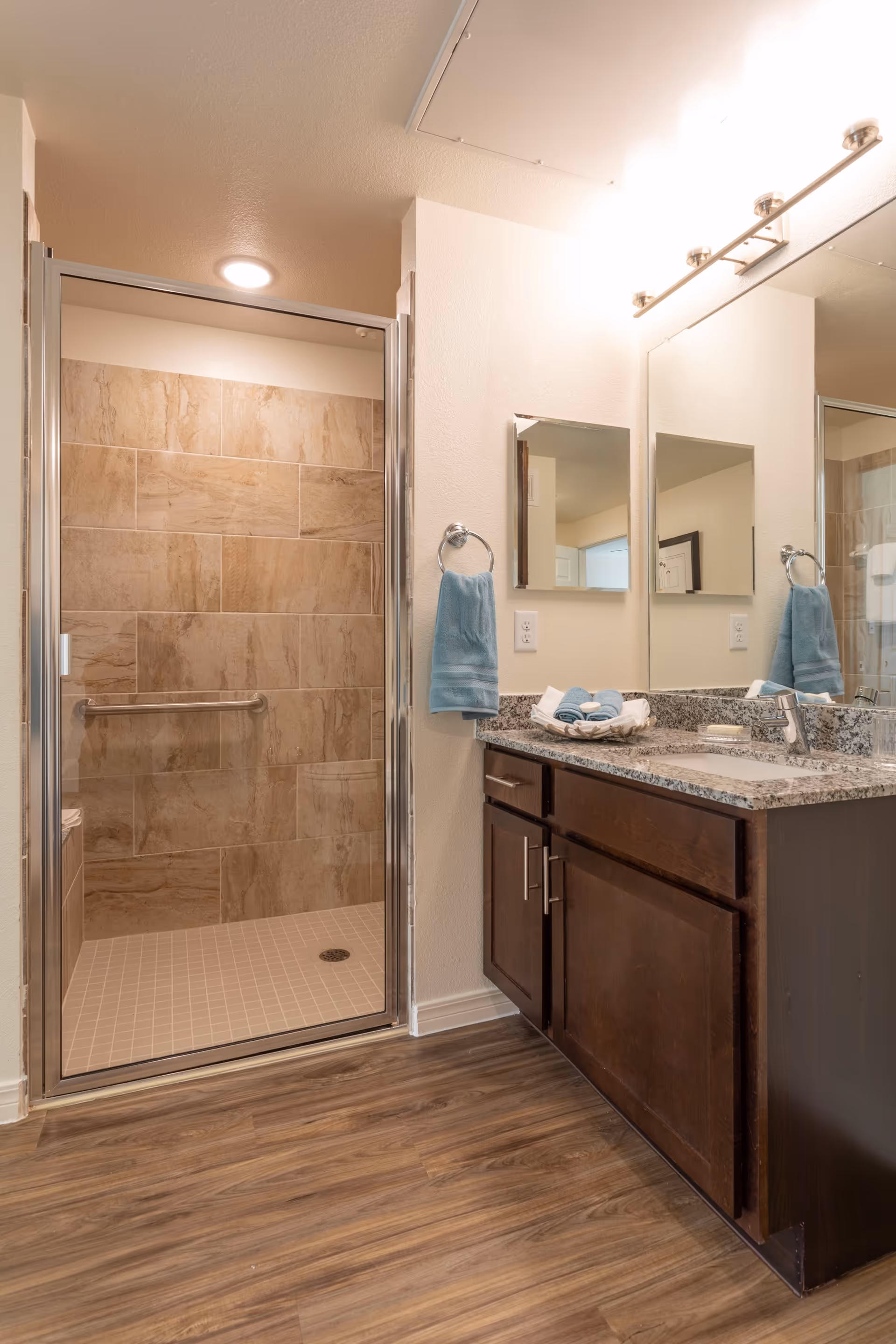 A modern bathroom featuring a walk-in shower with beige tiles and a glass door. Next to the shower is a vanity with a granite countertop, dark wood cabinets, a sink, and a large mirror above. Two blue hand towels hang on towel rings on either side of the vanity, and folded towels are placed on the countertop. The floor has wood-like vinyl planks.