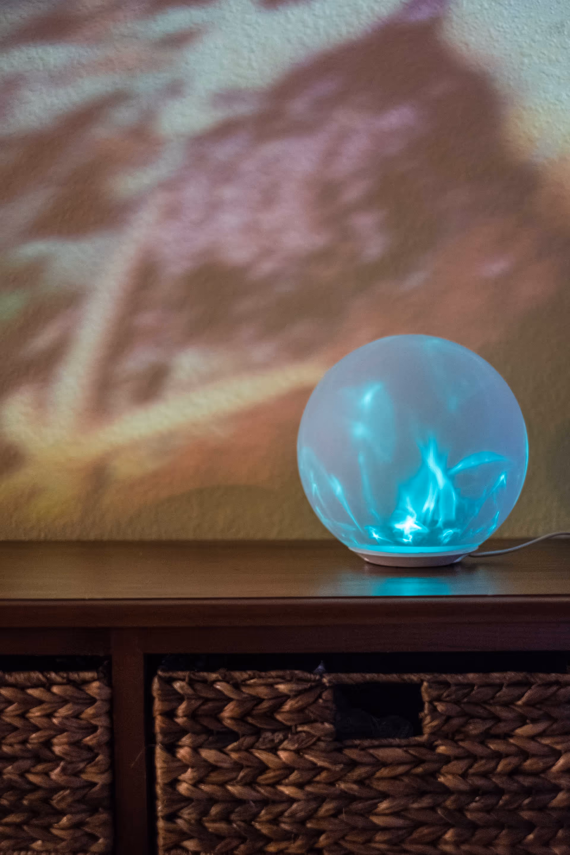 A glowing blue spherical lamp placed on a wooden surface with woven baskets underneath. The lamp casts colorful, abstract light patterns on the wall behind it.