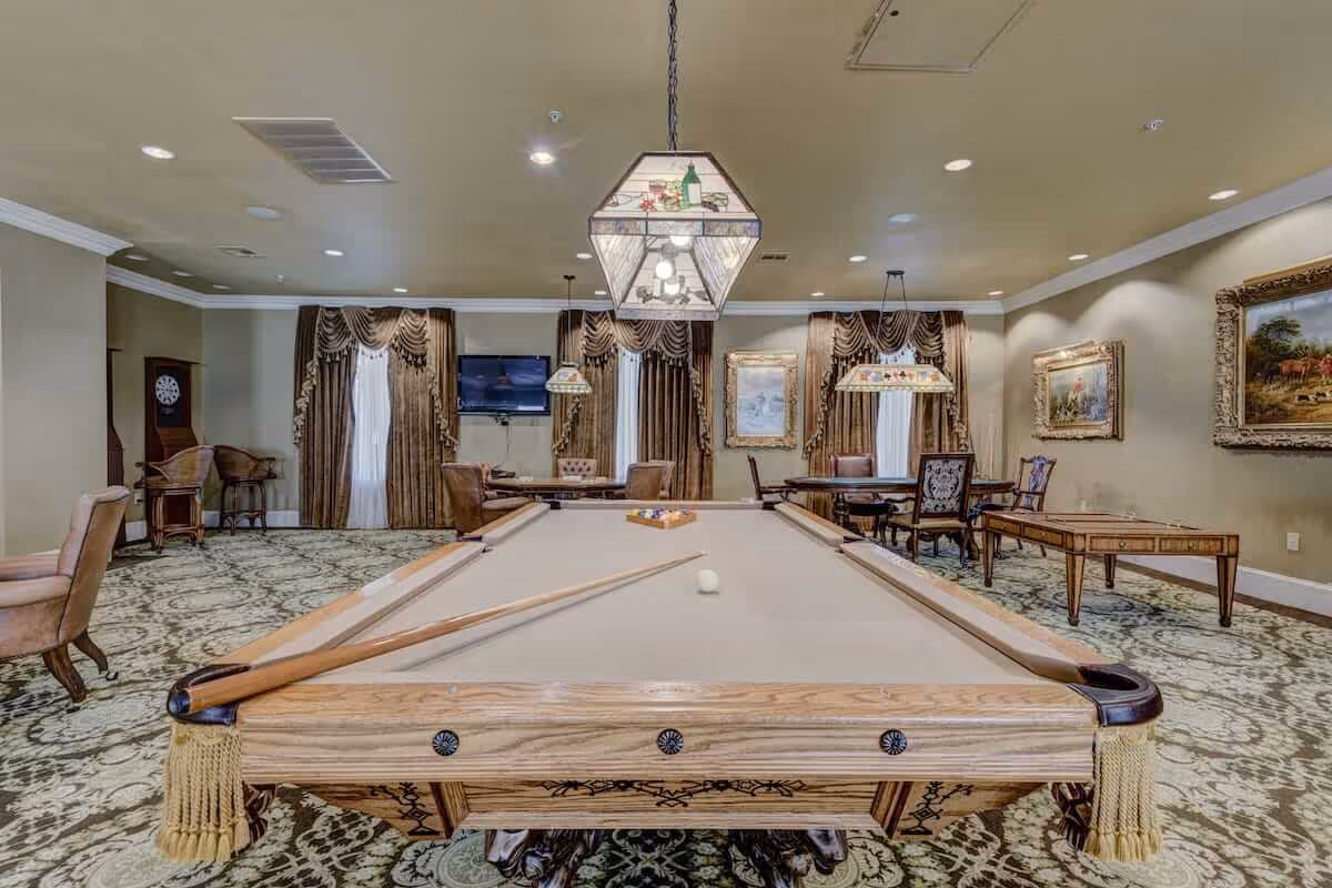 A spacious game room featuring a billiards table with cues and balls, surrounded by elegant chairs and tables. The room has patterned carpet, large windows with heavy drapes, framed paintings on the walls, and decorative hanging lights above the tables.