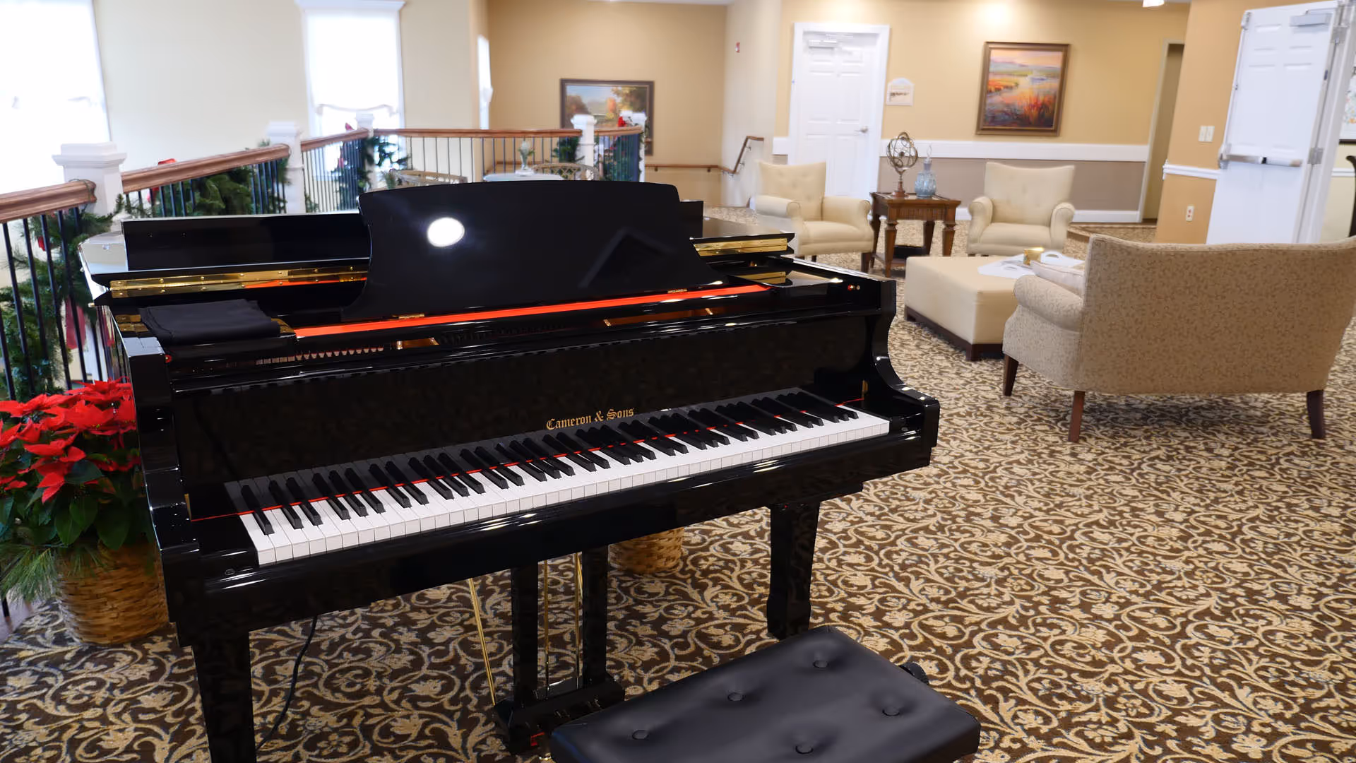 A black grand piano with a matching black bench is placed in a carpeted room with a patterned design. In the background, there are beige upholstered chairs and a matching ottoman arranged around a small wooden table. The room has cream-colored walls with white trim, a framed painting, and a whiteboard on the wall. There are also some potted red poinsettia plants near a railing decorated with greenery.