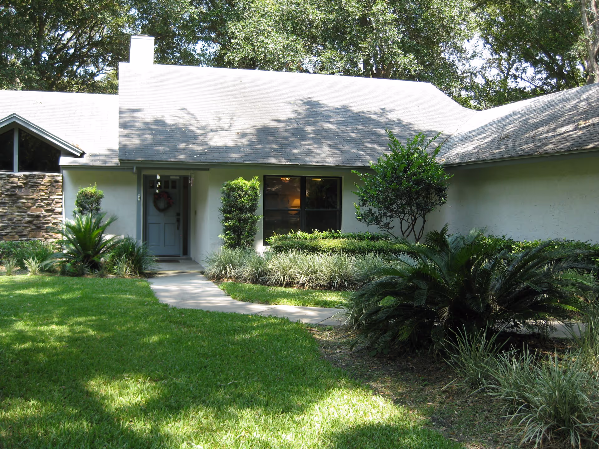 Front exterior view of a single-story house with a light gray roof and white walls, surrounded by green shrubs, plants, and a well-maintained lawn. A concrete pathway leads to a front door with a wreath hanging on it. Trees are visible in the background.