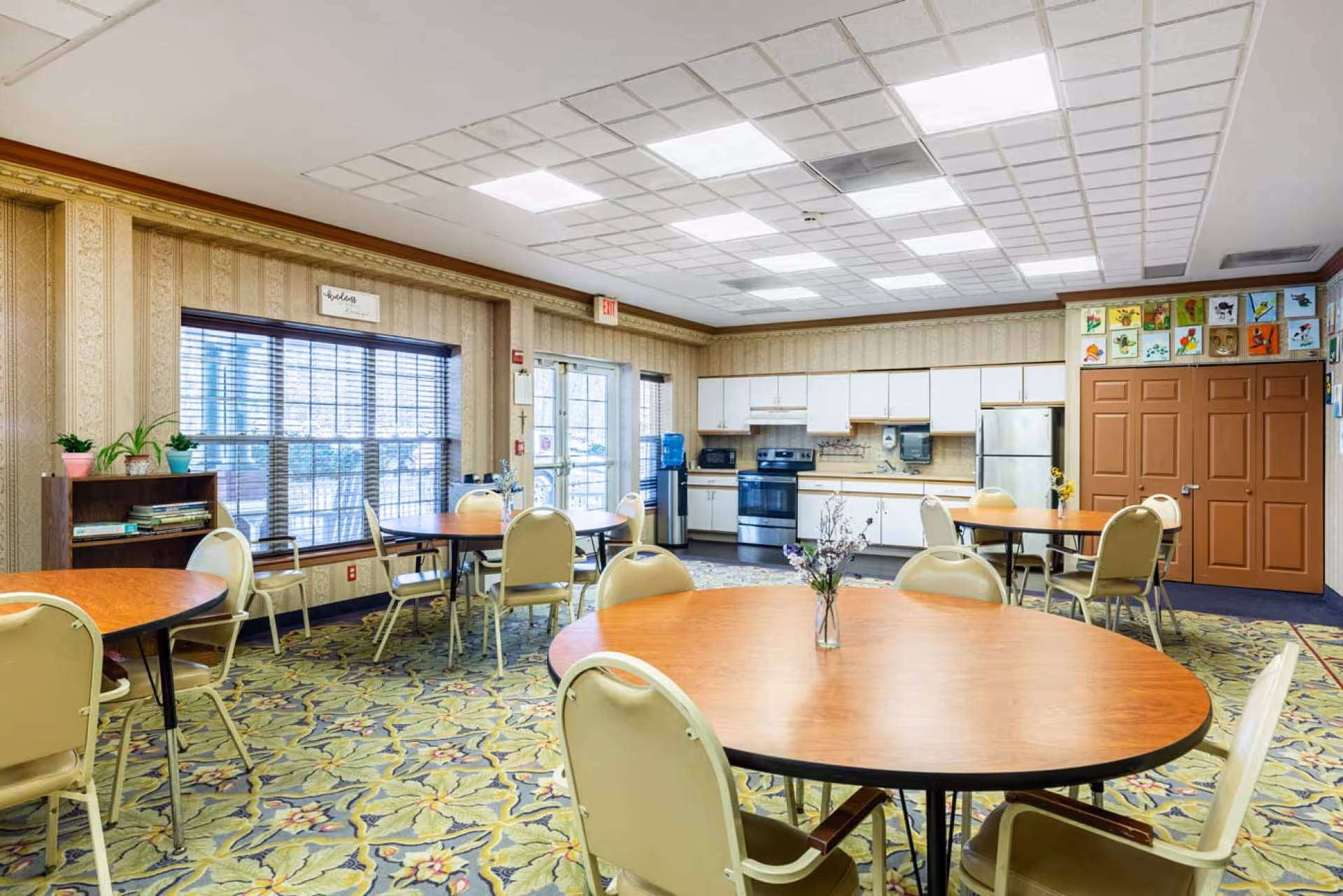 A bright and spacious dining room with round wooden tables and beige chairs. The room features a patterned carpet, large windows with blinds, and a kitchen area with white cabinets, a stove, microwave, and refrigerator. There are colorful drawings displayed above a set of brown double doors and a small bookshelf with potted plants near the window.