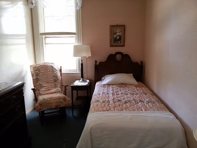 A small bedroom with a single bed covered with a patterned quilt and a white pillow. Next to the bed is a wooden nightstand with a lamp on it. There is a cushioned rocking chair with a floral pattern near a window with white curtains. A framed floral picture hangs on the wall above the bed.