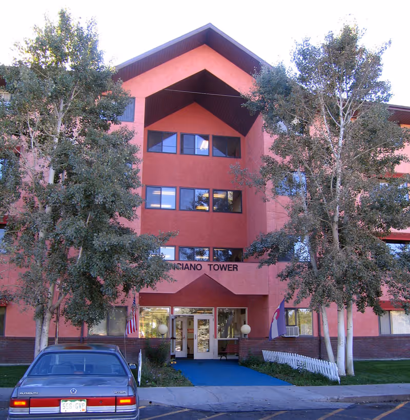 Front exterior of the Anciano Tower building showing the main entrance, flanking trees, and a parked car.