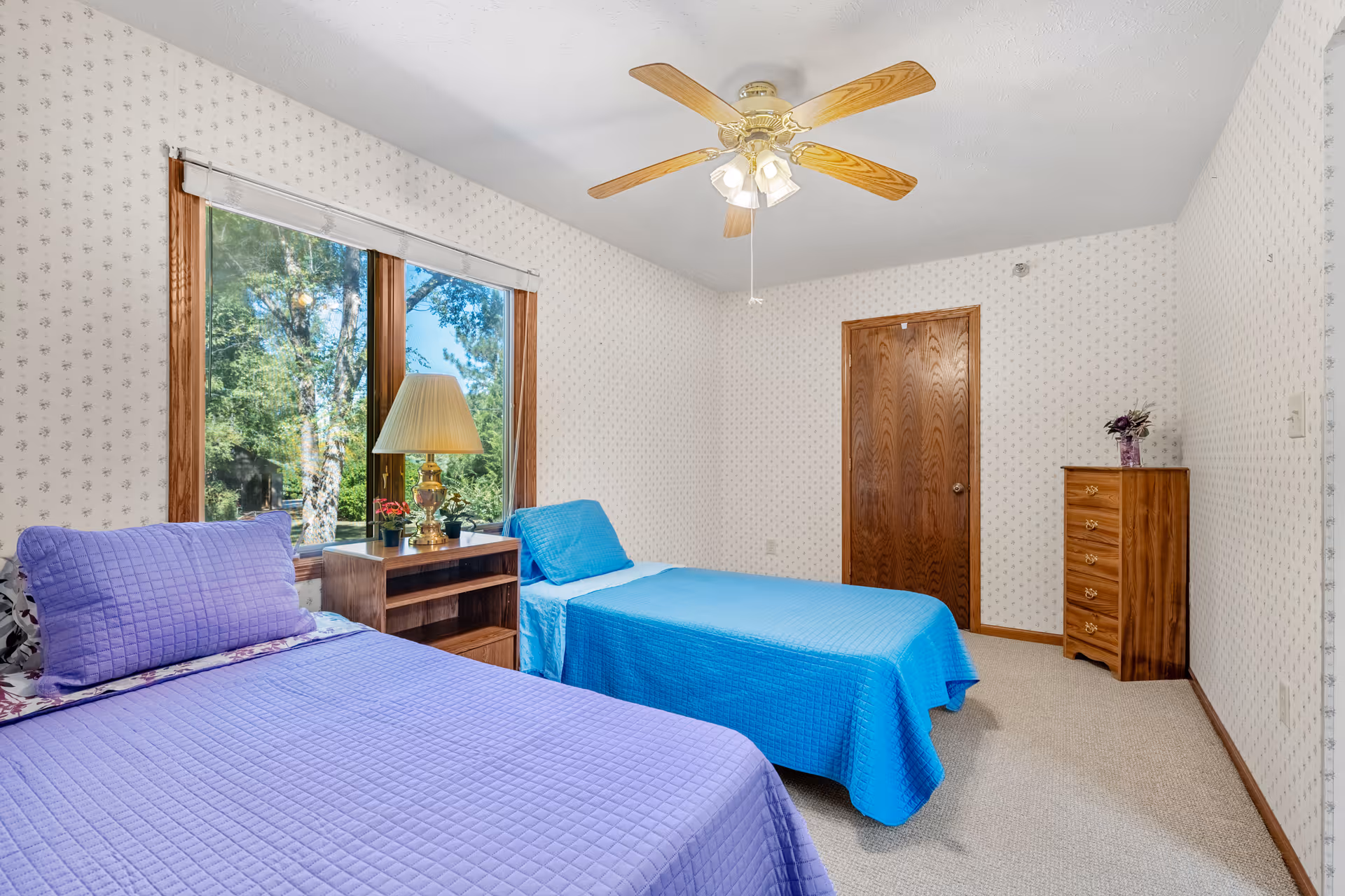 A bedroom with two single beds, one with a purple quilt and pillow and the other with a blue quilt and pillow. Between the beds is a wooden nightstand with a lamp and small potted plants. The room has a ceiling fan with lights, a wooden door, a wooden chest of drawers with a vase of flowers, and a large window showing green trees outside. The walls are covered with light-colored wallpaper with a subtle pattern.