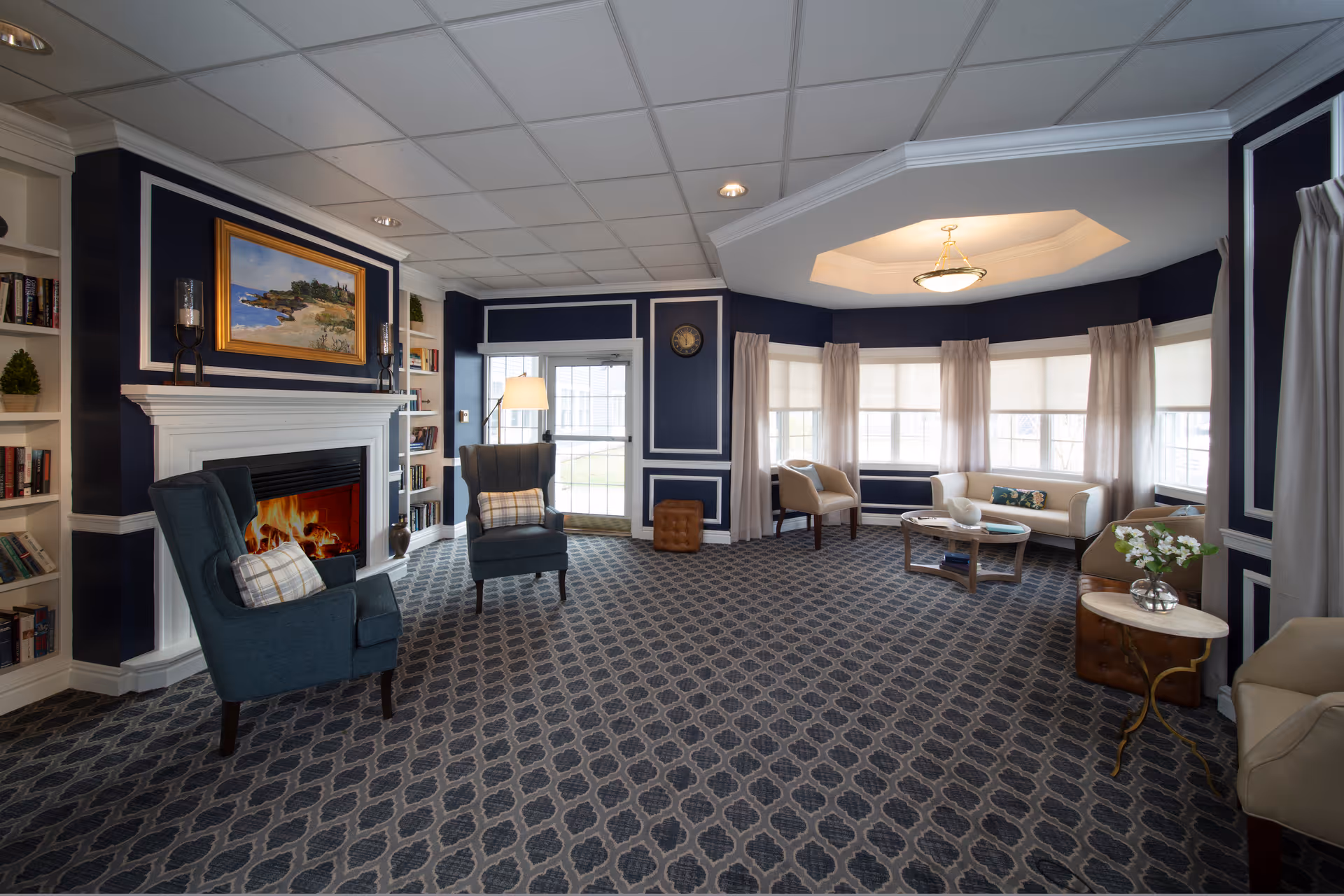 Spacious, well-lit common living room with upholstered chairs, a fireplace with built-in bookshelves, and a bay window seating area.