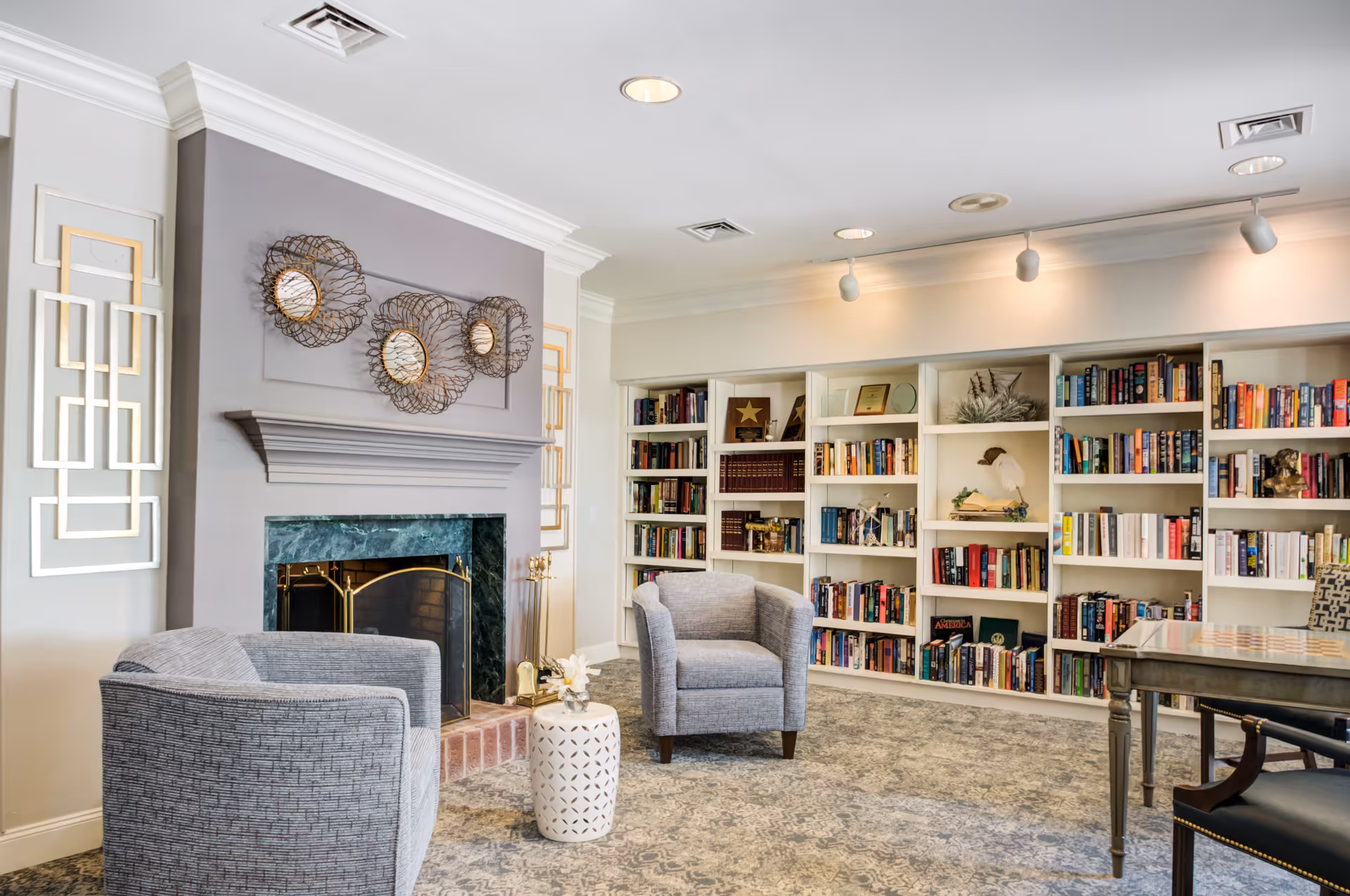 A cozy living room area featuring two gray upholstered armchairs positioned near a fireplace with a green marble surround and a gray mantel. Above the fireplace are three decorative circular metal wall art pieces with mirrors in the center. On either side of the fireplace are modern geometric wall decorations. The room has built-in white bookshelves filled with books and decorative items, and a table with chairs is partially visible on the right side. The ceiling has recessed lighting and track lights illuminating the space.
