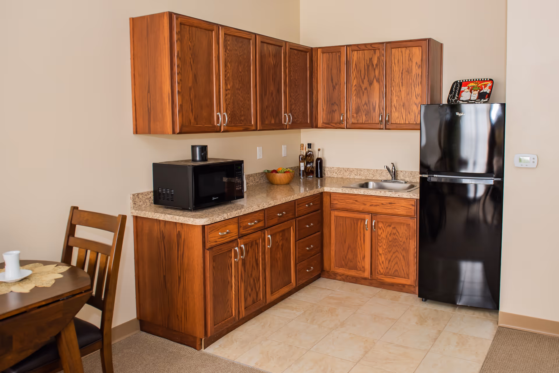 A small kitchen area with wooden cabinets, a black microwave on the countertop, a black refrigerator, a small sink, and a wooden dining table with chairs partially visible on the left side.