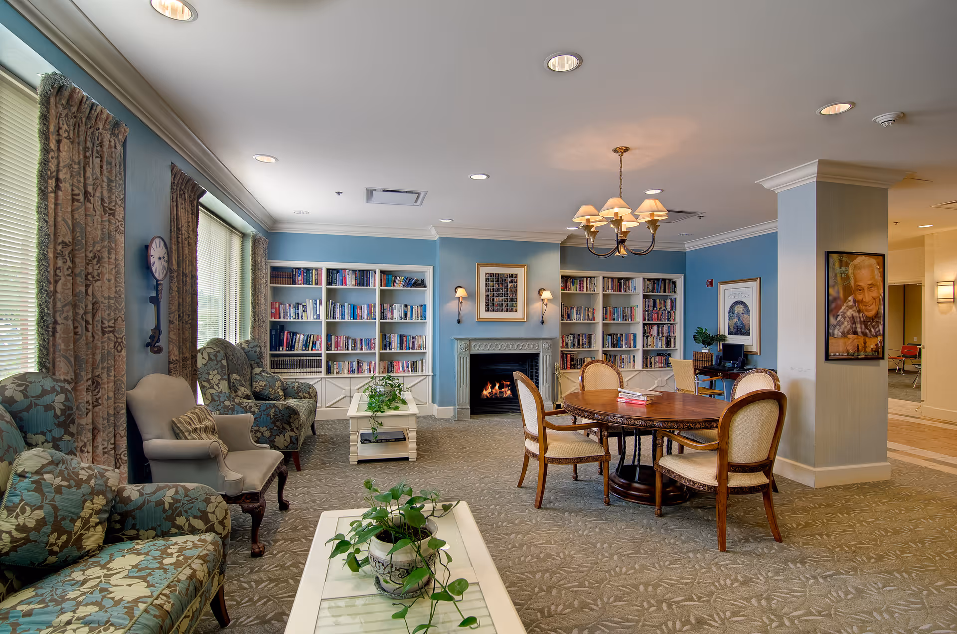 A cozy common area in a senior living facility with blue walls, a fireplace, built-in bookshelves filled with books, and a round wooden table with four chairs. There are several upholstered armchairs and a sofa with floral patterns, a coffee table with a potted plant, and large windows with patterned curtains letting in natural light. The ceiling has recessed lighting and a chandelier above the table. A framed portrait and artwork are visible on the walls.