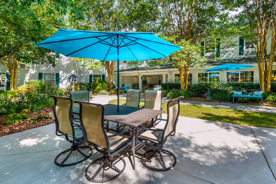 Outdoor patio area at Arbor Terrace Asheville featuring a rectangular table with six mesh chairs and a large blue umbrella providing shade. The patio is surrounded by greenery, trees, and a white building with windows and green shutters in the background. Another blue umbrella and bench are visible further back on the right side.