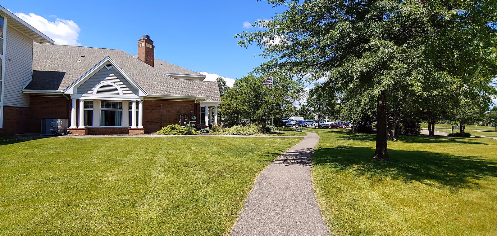 A paved walkway curves through a well-maintained green lawn with trees on the right side, leading towards a brick and siding building with white columns and a chimney under a clear blue sky.
