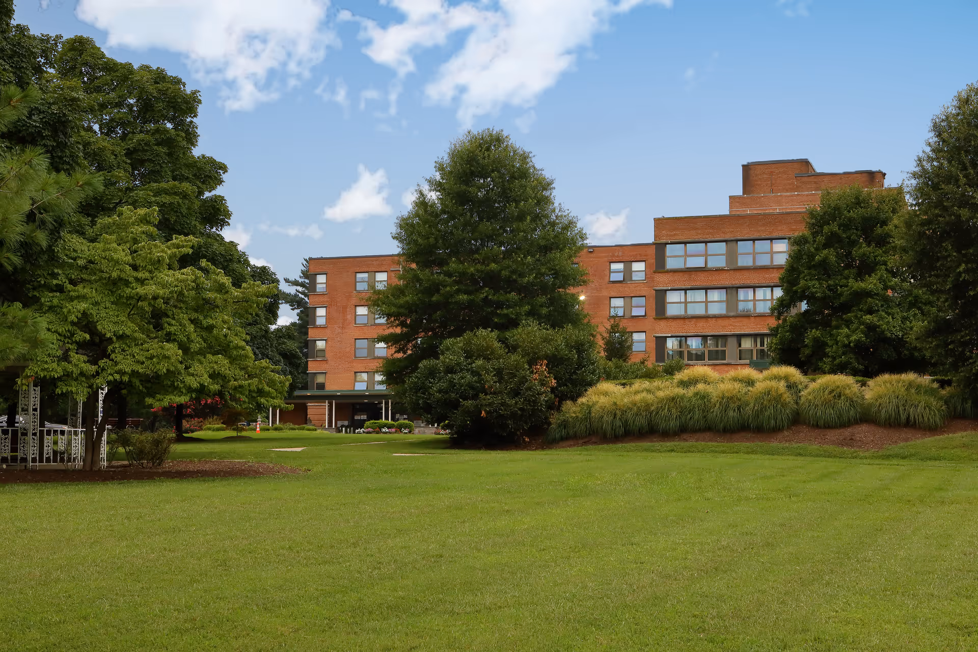 A large brick building with multiple windows surrounded by green trees and bushes under a partly cloudy blue sky. In the foreground, there is a well-maintained grassy lawn with landscaped shrubs and trees.