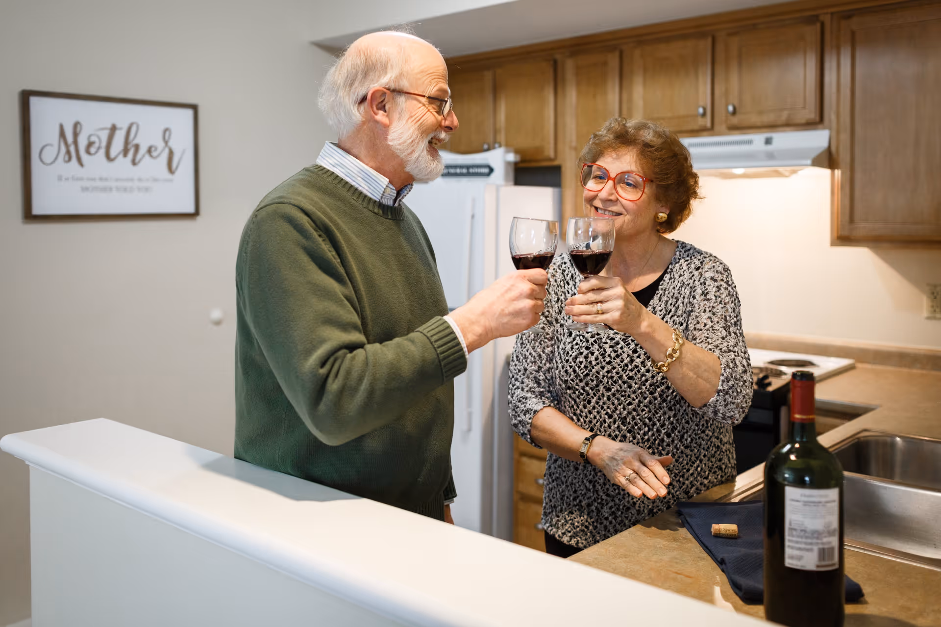 An elderly man and woman clink wine glasses in a kitchen. The man is wearing a green sweater and glasses, and the woman is wearing a patterned top and red glasses. A bottle of wine and a cork are on the kitchen counter. Wooden cabinets and a refrigerator are visible in the background. A framed sign on the wall reads 'Mother'.