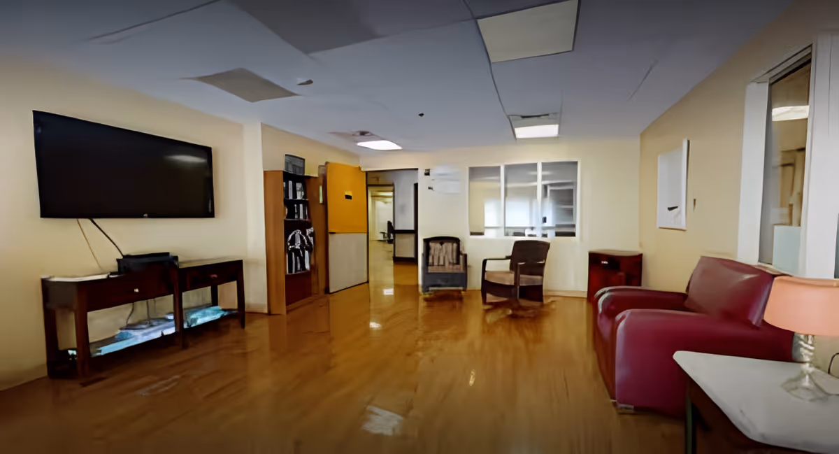 A waiting or common area with wooden flooring, a large flat-screen TV mounted on the wall, a wooden console table beneath the TV, two armchairs, a red sofa, a small side table with a lamp, and a bookshelf. The ceiling has some visible damage and missing panels.