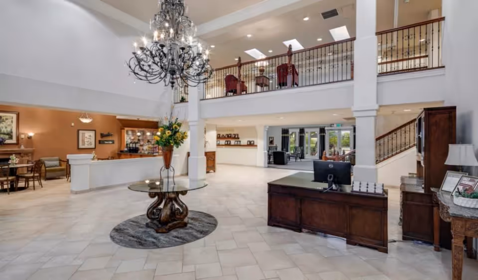 Spacious and elegant senior living facility lobby with a large chandelier hanging from the ceiling, a round glass table with a floral arrangement in the center, a wooden reception desk with a computer, seating areas with chairs and tables, and a staircase leading to an upper balcony with additional seating.