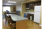 Communal dining and kitchenette area with a long island table surrounded by chairs, cabinets, stove and refrigerator.