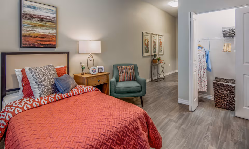 A cozy bedroom with a bed covered in a red quilt and multiple pillows, a wooden nightstand with a lamp and decorative items, a green armchair with a striped pillow, and an open closet with clothes hanging and woven baskets inside. The room has light-colored walls, wood flooring, and framed artwork on the walls.